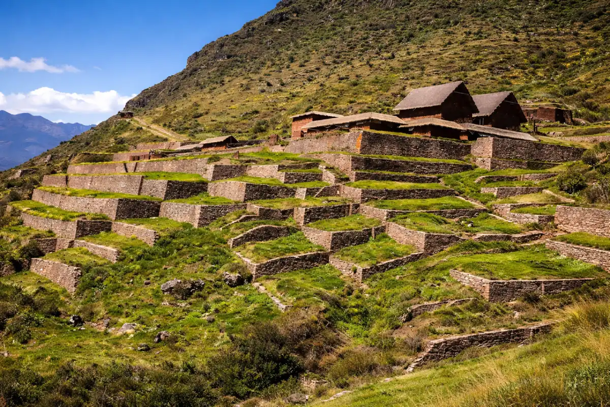 Vista de terrazas de piedra en Huchuy Qosqo, con estructuras andinas sobre una ladera montañosa cubierta de vegetación bajo un cielo despejado.