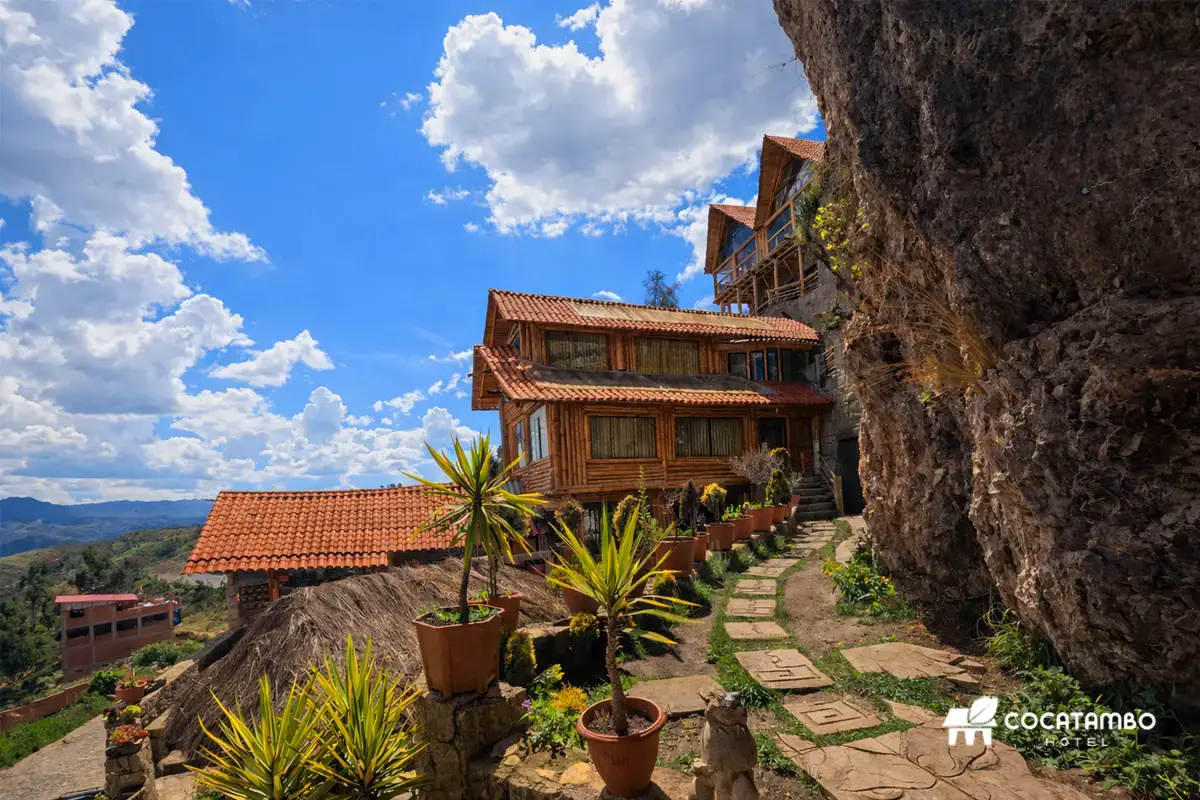 Camino de piedra junto a un edificio rústico de madera en ladera rocosa con cielo azul y nubes.