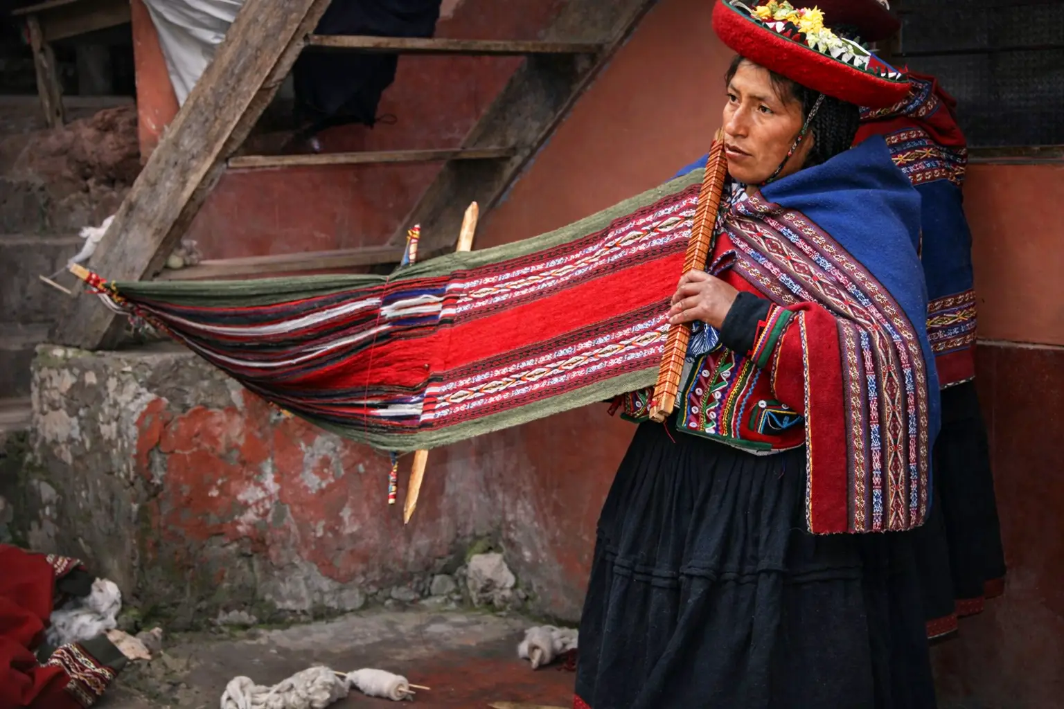 Artesana de Chinchero tejiendo en telar de cintura con manto rojo y azul, frente a una pared de adobe.