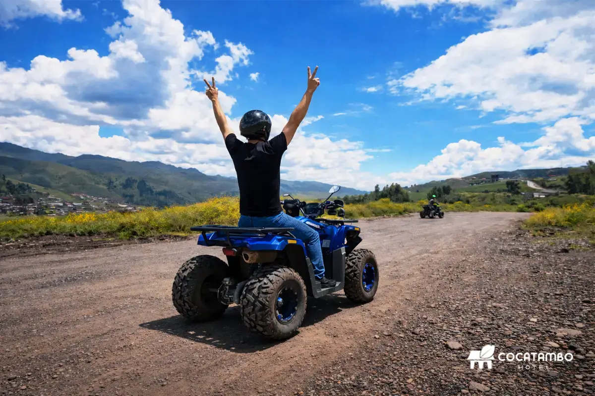 Persona en cuatrimoto azul con los brazos levantados sobre camino de tierra, con montañas y cielo abierto al fondo.