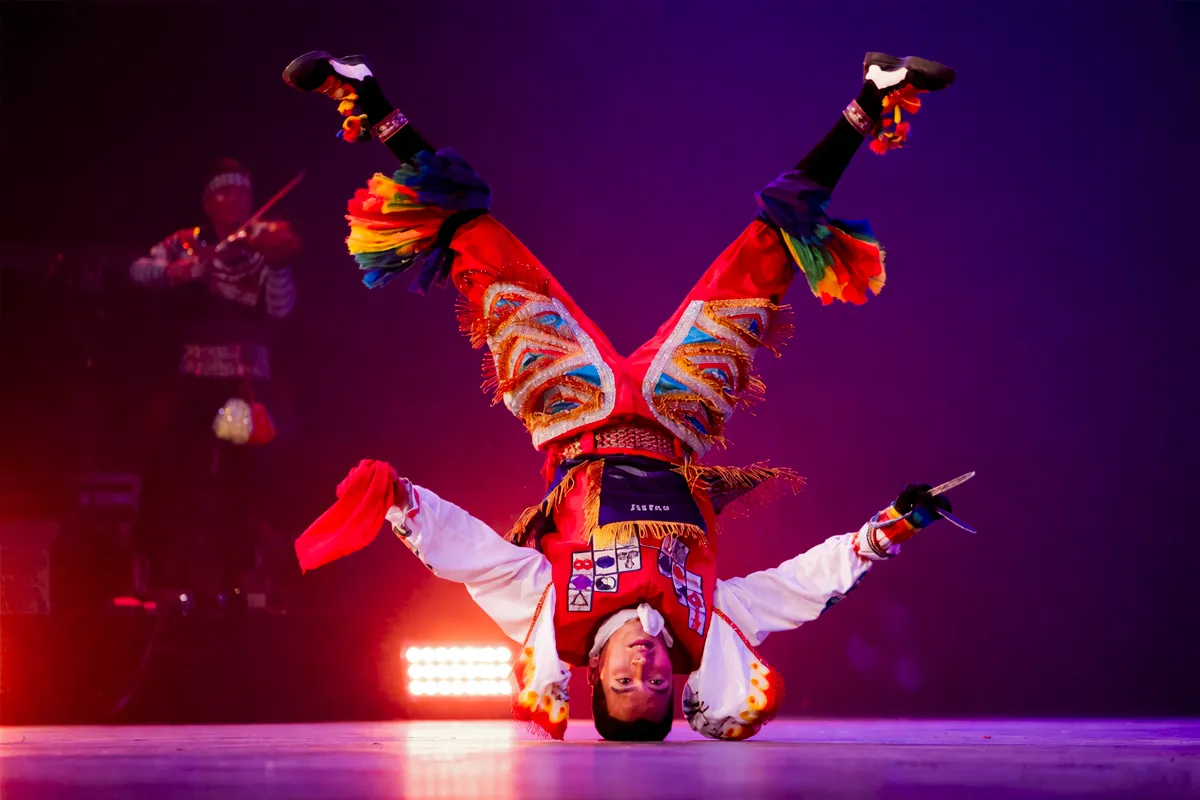 Danzante de tiejeras realizando acrobacia invertida en escenario iluminado, con traje rojo tradicional y músico al fondo.