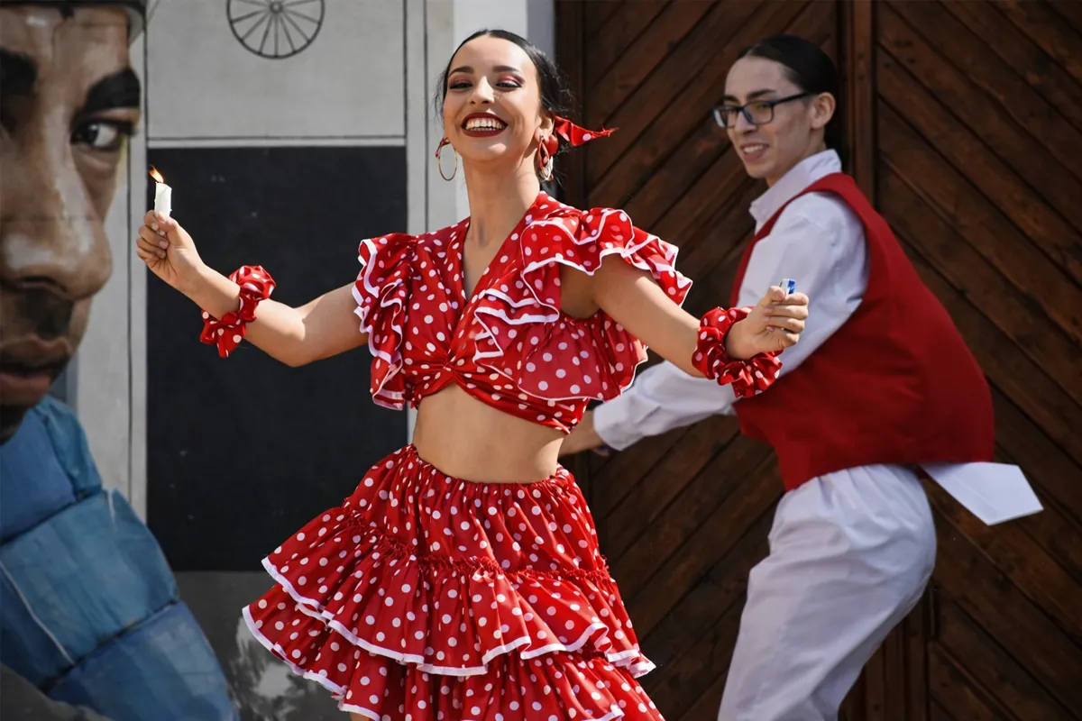 Bailarina de festejo con vestido rojo de lunares sonriendo durante una presentación callejera, acompañada por otro artista al fondo.