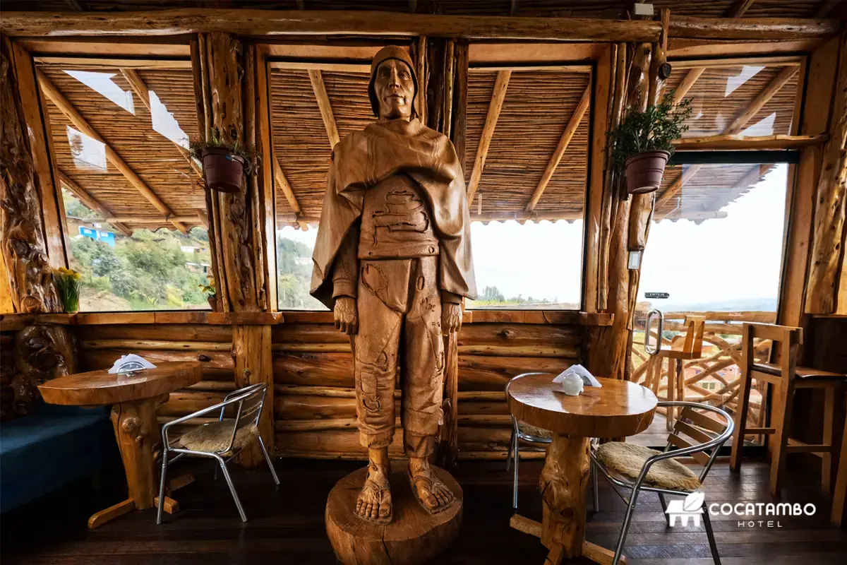Escultura de madera del Gigante de Paruro en el interior de un restaurante rústico con mesas, sillas y grandes ventanales.
