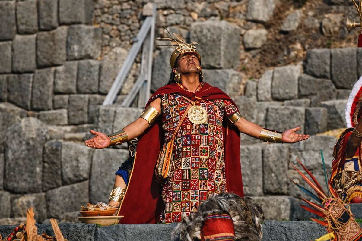 Representación del Inca durante una ceremonia en Sacsayhuamán, con los brazos abiertos, vestido con atuendo ceremonial rojo y dorado, frente a muros de piedra incas.