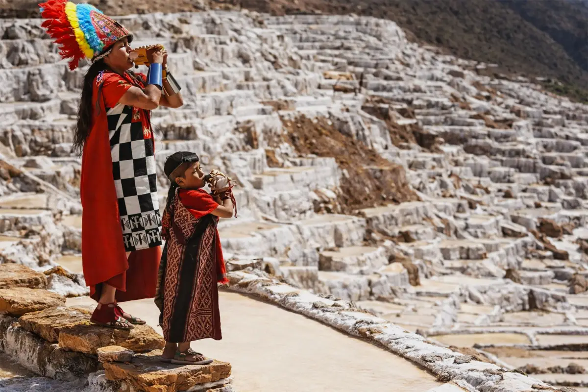 Personas con vestimenta tradicional andina tocando instrumentos frente a las terrazas de las Salineras de Maras