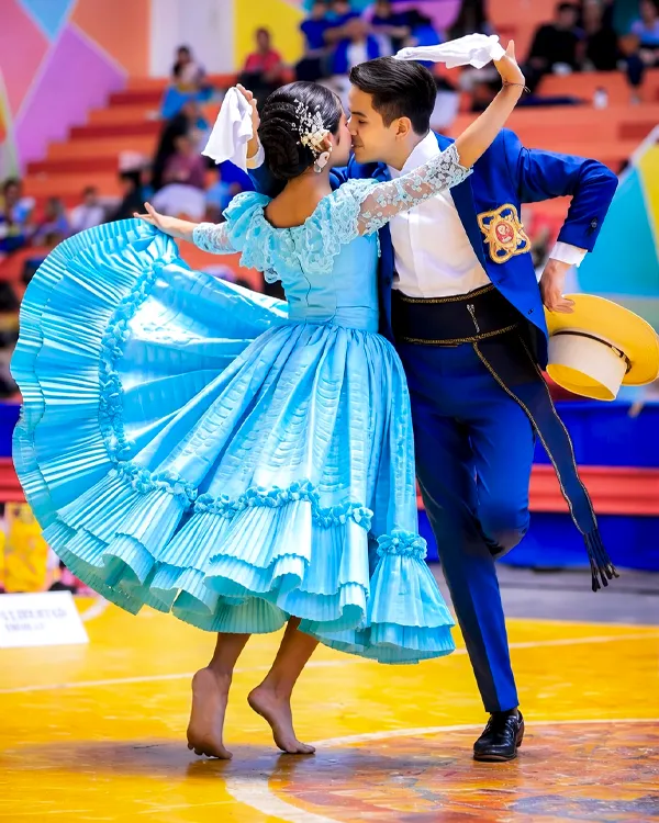 Pareja bailando marinera en escenario interior, con vestido celeste amplio y traje azul, en plena presentación folclórica.