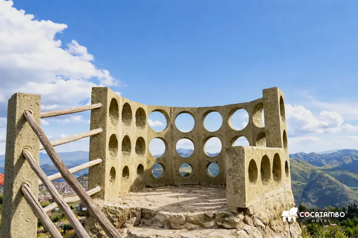 Mirador de piedra con muros perforados circulares y vista abierta a las montañas bajo cielo azul.