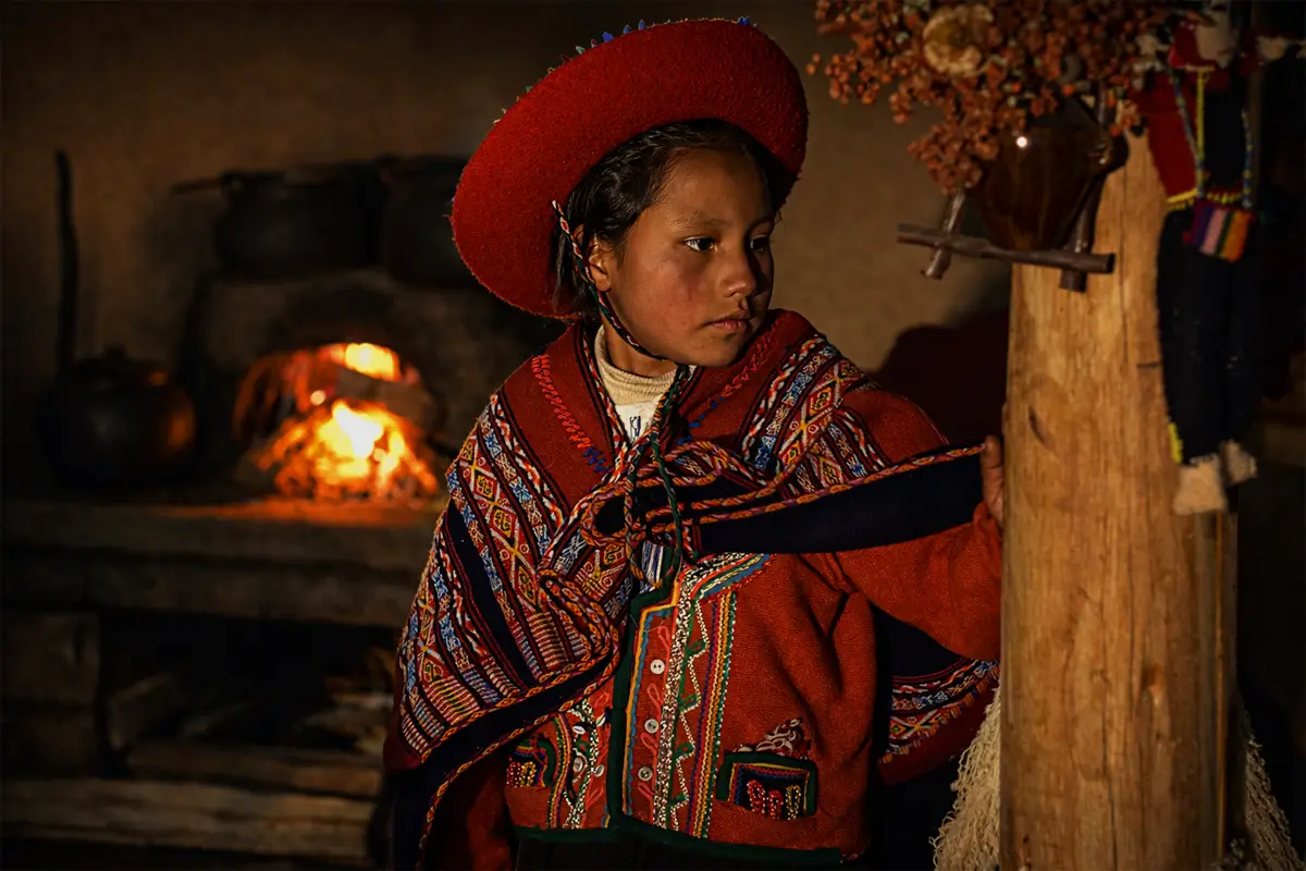 Niña de Chinchero con vestimenta tradicional andina, junto a un fogón dentro de una cocina rústica.