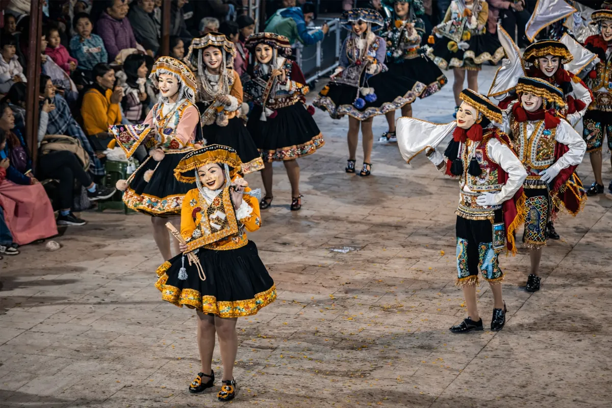 Grupo de bailarines de Paucartampus con trajes bordados y sombreros blancos presentándose en avenida urbana durante fiesta tradicional.