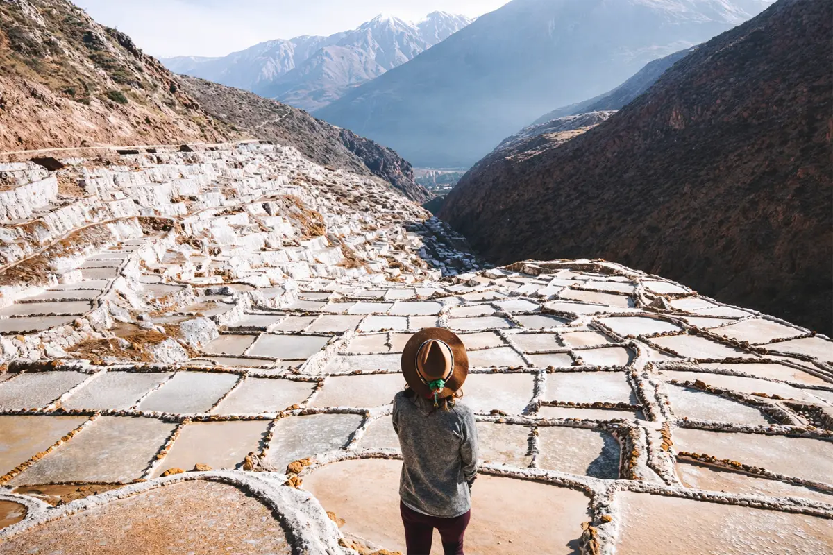 Viajera con sombrero observando las Salineras de Maras desde un mirador, con terrazas de sal y montanas al fondo