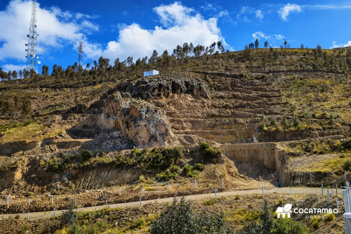 Vista de una cantera en ladera andina con terrazas de excavación, camino de tierra, torres de comunicación y cielo azul con nubes.