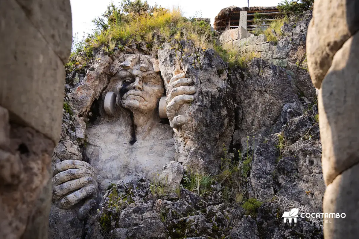 Escultura de Tocay Capac tallada en la roca, vista entre muros de piedra, con vegetación alrededor.