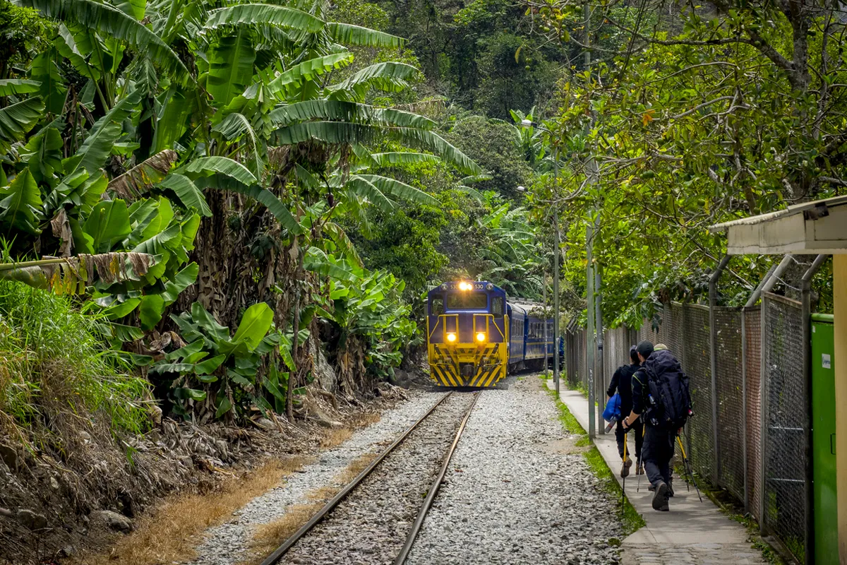 Tren acercándose por la vía férrea cerca de Machu Picchu mientras viajeros caminan junto a los rieles