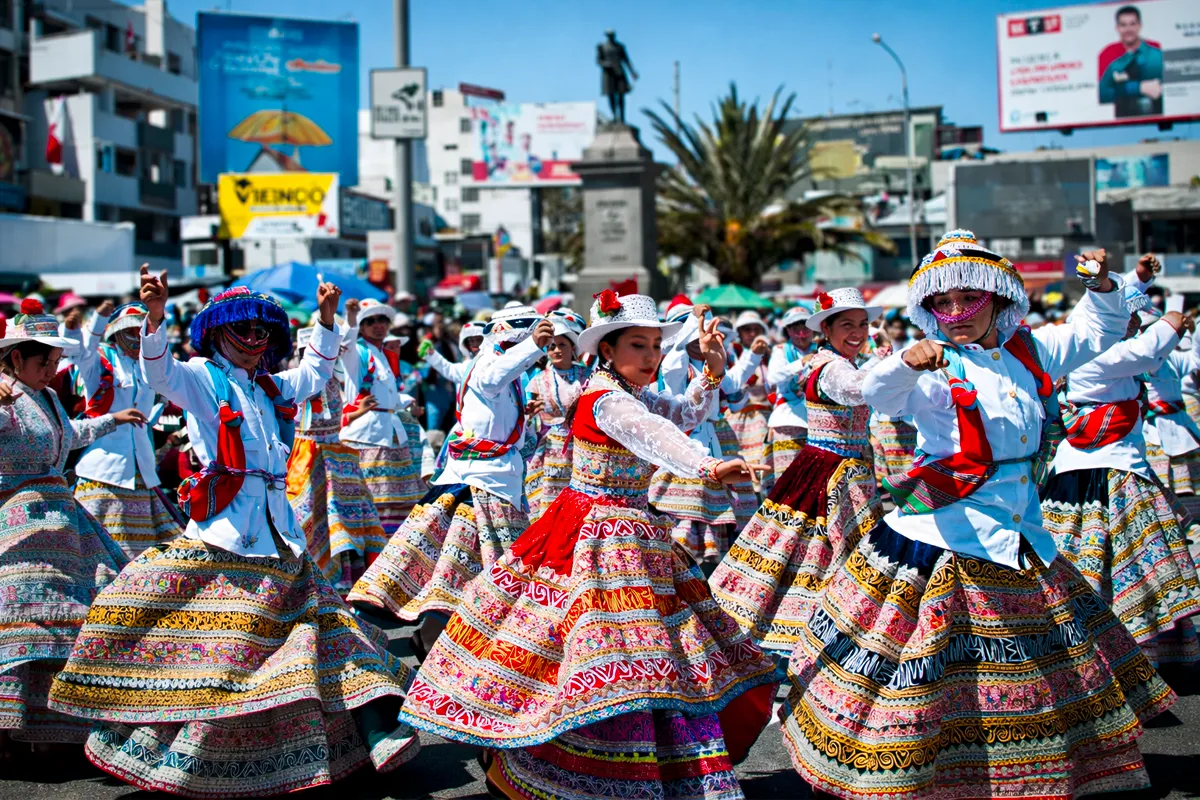 Grupo de Danzantes de Wititi con trajes bordados y sombreros blancos presentándose en avenida urbana durante fiesta tradicional.